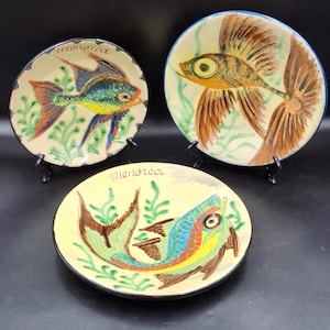 Puede incluir: Tres platos redondos de cerámica con diseños de peces pintados a mano en tonos azul, amarillo y marrón, con detalles de algas verdes. Cada plato muestra la palabra Menorca escrita en cursiva en el borde superior de la superficie crema.