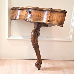 May include: A vintage, ornate wooden side table with a single drawer and a carved leg. The table is a warm brown color, with visible wood grain. The drawer has a small, round knob. The table is approximately 30 inches wide.