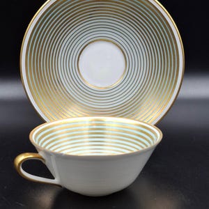 May include: White teacup and saucer set with gold and light blue concentric stripes. The teacup has a gold handle and the saucer has a matching design. The set is on a black surface.