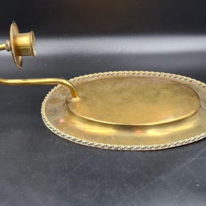 Puede incluir: Un candelero de latón vintage con base ovalada y brazo curvo. La base tiene un borde decorativo en forma de cuerda. El receptáculo de la vela es circular. El latón tiene un tono dorado cálido, lo que sugiere antigüedad. El diseño es simple pero elegante.