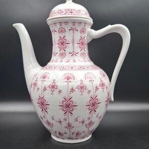 May include: White ceramic coffee pot with a lid and handle. The pot is decorated with a repeating pattern of pink floral designs. The spout is long and curved. The handle is a curved, open design. The lid has a small knob on top.