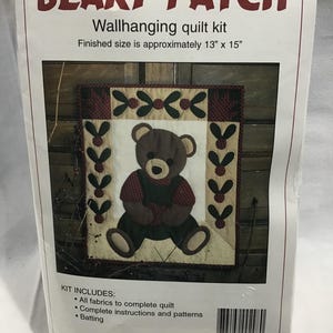 May include: A wallhanging quilt kit featuring a brown teddy bear with a red and white plaid shirt. The kit includes all fabrics, instructions, and patterns to complete a quilt measuring 13 inches by 15 inches. The kit also includes batting.