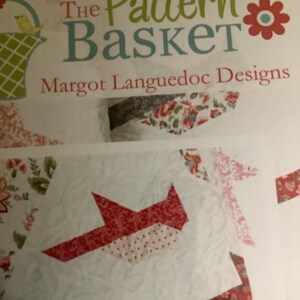 May include: The Pattern Basket by Margot Languedoc Designs. The image shows a quilt pattern with red and white fabric pieces arranged in a geometric design. The quilt has a floral pattern.