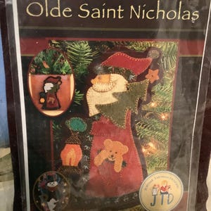 May include: A craft kit for a seasonal wall pocket, featuring a design of Olde Saint Nicholas. The kit includes felt cutouts of Santa Claus, a teddy bear, and other Christmas-themed ornaments. The text "Olde Saint Nicholas" is displayed at the top.