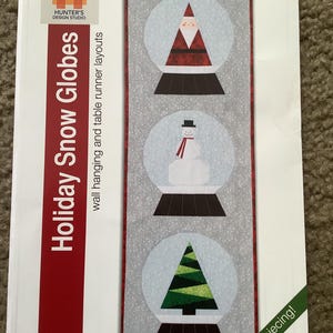 May include: A fabric wall hanging or table runner featuring three snow globes with holiday designs. The snow globes contain a Santa, a snowman, and a Christmas tree. The text "Holiday Snow Globes" is visible.