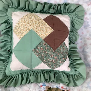 May include: Square decorative pillow with a white center and a patchwork design featuring floral and solid fabrics in shades of green, brown, and yellow. The pillow is framed by a ruffled green border.