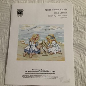 May include: A printed cross-stitch pattern titled "Sand Castles" by Linda Gillum. The design features two children building a sandcastle on a beach, with seagulls in the sky. The pattern is from Kooler Classic Charts.