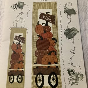 May include: A fabric panel featuring a stack of pumpkins in a wagon, with the word "Fall" on a sign. The panel is 6 inches by 22 inches. The design includes various textures and colors of orange, brown, and tan.