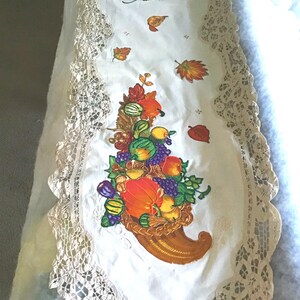 May include: White table runner with a lace border and an embroidered cornucopia filled with fall fruits and vegetables. The cornucopia is surrounded by leaves and pumpkins.