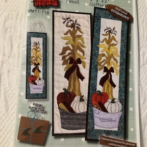 May include: A craft pattern for front door decor, featuring a fall harvest theme. The design includes corn stalks, pumpkins, and a bucket. The pattern is available in two sizes: 6" x 18" or 6" x 22". Includes a button pack.