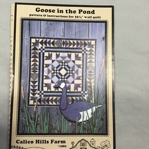 May include: A quilt pattern for a 35 1/2 inch wall quilt titled "Goose in the Pond" by Calico Hills Farm. The pattern features a blue goose in a pond with reeds and a wooden fence in the background. The quilt pattern is from 1982.