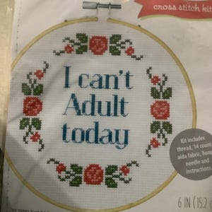 May include: Cross stitch kit with the text "I can't Adult today" in teal. The design includes floral accents in red and green. The kit includes thread, aida fabric, a hoop, a needle, and instructions. The kit is 6 inches (15.2 cm).