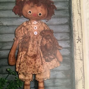 May include: A rag doll with red yarn hair, a brown dress with white buttons, and a brown teddy bear in her arms. The doll is standing in front of a wooden wall.