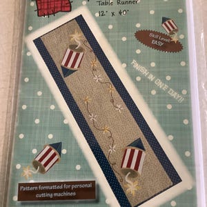 May include: A table runner kit with a patriotic theme. The design features red, white, and blue fireworks and floral accents. The text on the package reads "Spark of Fun" and "12" x 40"", indicating the runner's dimensions. The skill level is "EASY".