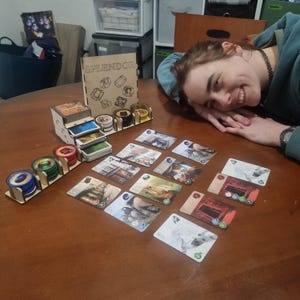 Może przedstawiać: Drewniany organizer do gry planszowej Splendor, z miejscami na karty, uchwytami na żetony i pudełkiem z tytułem gry. Karty i żetony gry są ułożone na brązowym stole. Obok gry uśmiecha się osoba.