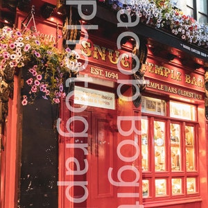 May include: A red brick building with a sign that reads "Snug Temple Bar Est. 1694". The building has a large red door and a window display with various items inside. The building is decorated with hanging flower baskets.