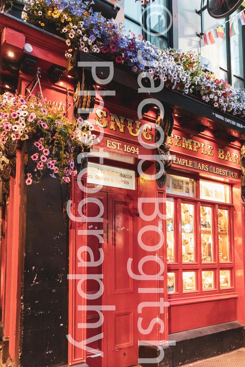 May include: A red brick building with a sign that reads "Snug Temple Bar Est. 1694". The building has a large red door and a window display with various items inside. The building is decorated with hanging flower baskets.