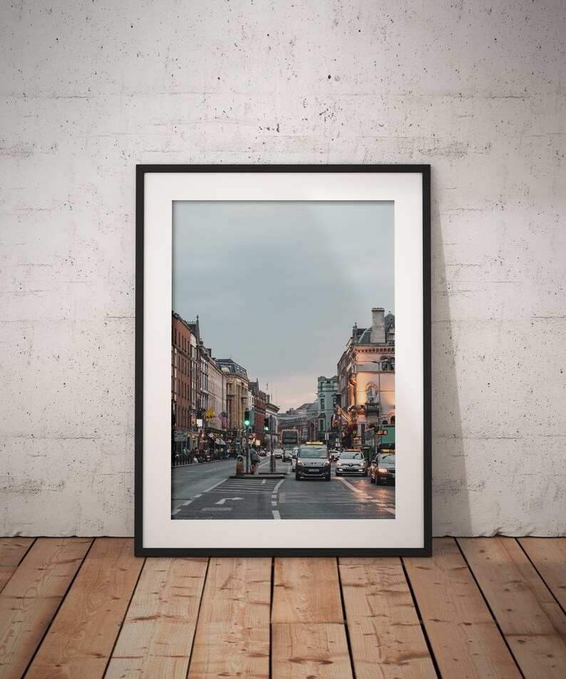 May include: A framed photograph of a city street with buildings on either side. The street is lined with cars and there are traffic lights in the distance. The sky is overcast and the buildings are a mix of brick and stone.