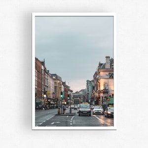 May include: A street scene in a city with buildings on both sides of the street. There are cars and a bus driving down the street. The sky is overcast and the buildings are a mix of brick and stone.
