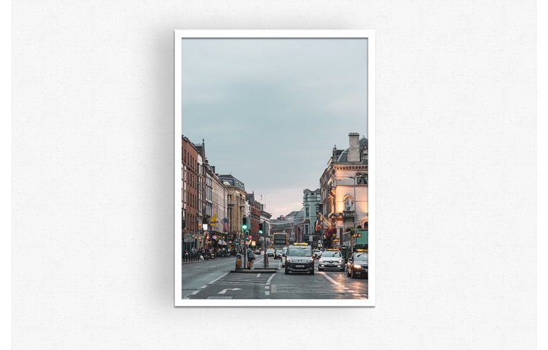 May include: A street scene in a city with buildings on both sides of the street. There are cars and a bus driving down the street. The sky is overcast and the buildings are a mix of brick and stone.