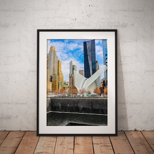 Puede incluir: Una fotografía enmarcada del Memorial y Museo del 11-S en la ciudad de Nueva York. La foto muestra las piscinas reflectantes y los rascacielos circundantes.