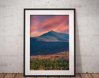 May include: Framed landscape print featuring a mountain peak under a vibrant sunset sky. The image showcases a forest with autumn foliage in shades of green, yellow, and orange. The print is displayed in a black frame, leaning against a white wall on a wooden floor.