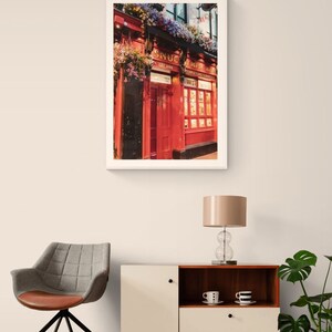 May include: A framed photograph of a red brick building with a sign that reads "The Snug Empire Bar" and a red door. The building is decorated with hanging flower baskets.