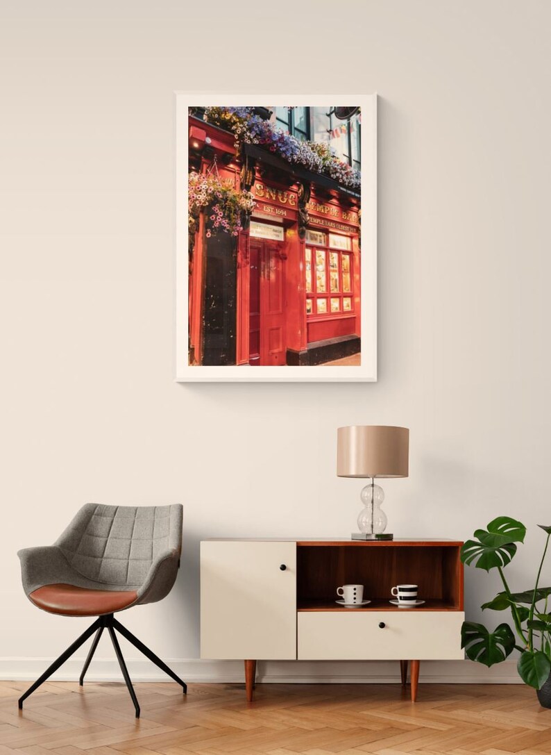 May include: A framed photograph of a red brick building with a sign that reads "The Snug Empire Bar" and a red door. The building is decorated with hanging flower baskets.