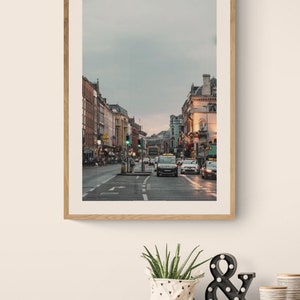 May include: A framed photograph of a city street with buildings on both sides and cars driving down the street. The sky is overcast and the street is wet.