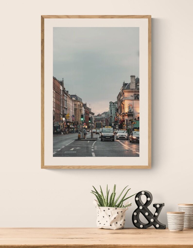 May include: A framed photograph of a city street with buildings on both sides and cars driving down the street. The sky is overcast and the street is wet.