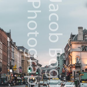 May include: A street scene in a city with tall buildings on either side of the road. There are cars driving down the road, including a yellow taxi. The sky is overcast and grey.