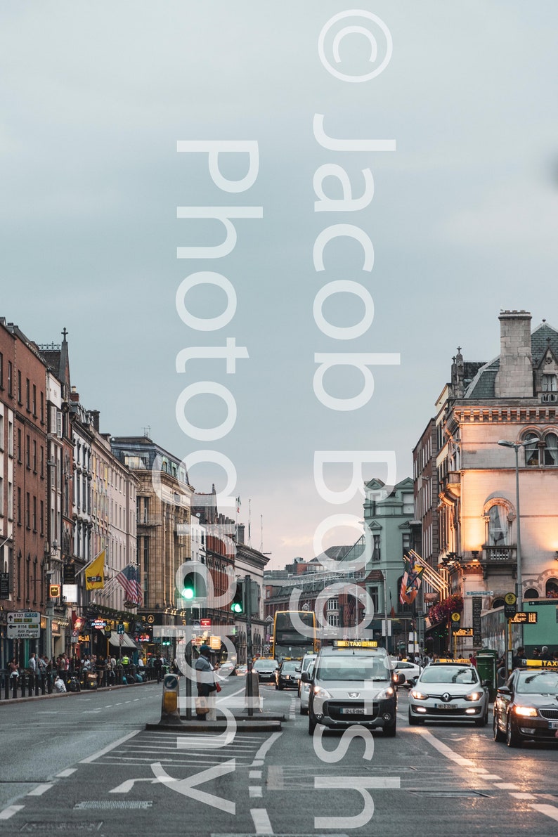 May include: A street scene in a city with tall buildings on either side of the road. There are cars driving down the road, including a yellow taxi. The sky is overcast and grey.