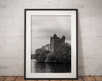 May include: Black and white photograph of a stone castle on a small island in a lake. The castle is surrounded by trees and the sky is cloudy.