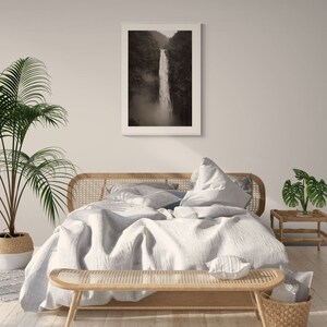 May include: A white bed with a light gray duvet and a woven headboard. The bed is styled with a white throw pillow and a woven bench at the foot of the bed. A black and white photograph of a waterfall hangs on the wall above the bed.