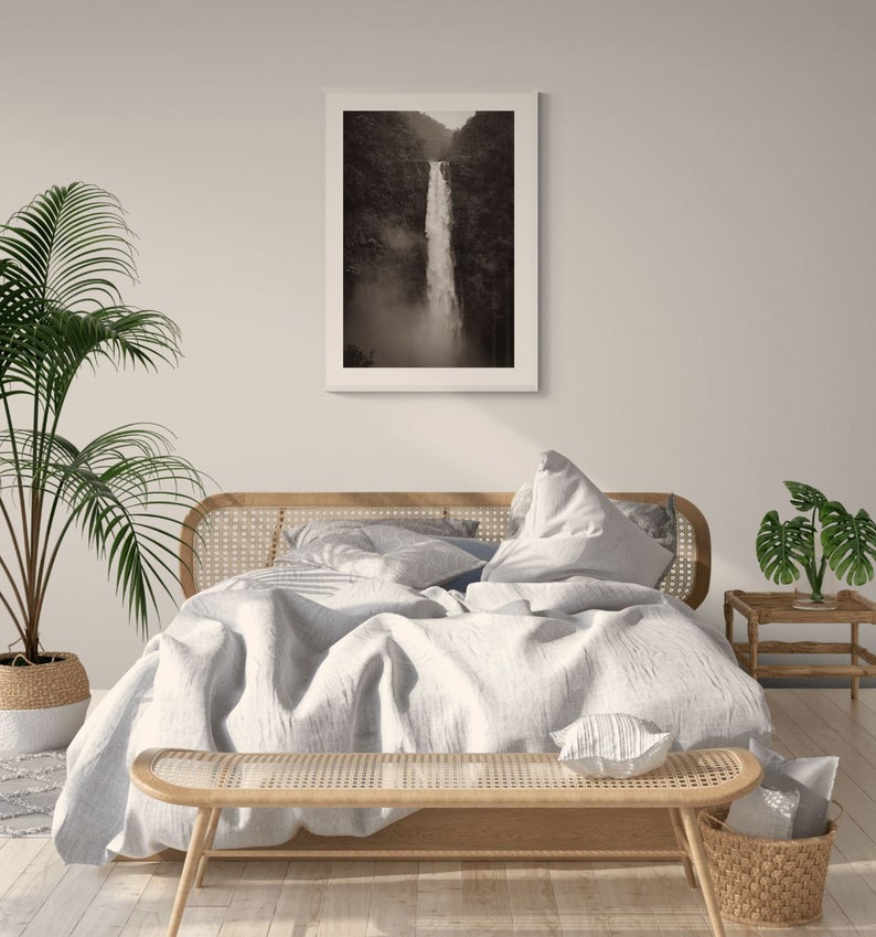 May include: A white bed with a light gray duvet and a woven headboard. The bed is styled with a white throw pillow and a woven bench at the foot of the bed. A black and white photograph of a waterfall hangs on the wall above the bed.