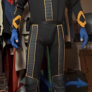 May include: A black full-body jumpsuit featuring yellow stitched accents, a grey circular emblem on the chest, blue gloves, and a blue tail. The suit includes ribbed detailing on the torso and legs, with yellow trim along the arms and thighs.