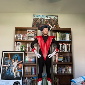 Könnte beinhalten: Eine Person in einem schwarzen und roten Kostüm mit einem V-förmigen Bruststück. Die Person steht vor einem Bücherregal mit einem Star Wars-Poster links.