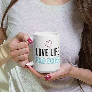 Könnte beinhalten: Weiße Keramiktasse mit einem schwarzen und rosafarbenen Herzdesign. Die Tasse trägt den Schriftzug "LOVE LIFE read BOOKS" in Schwarz und Blau.