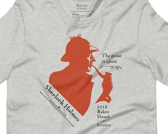Pode incluir: Uma camiseta cinza com uma silhueta vermelha de Sherlock Holmes, um cachimbo e o texto "The game is afoot." O texto "Sherlock Holmes" está escrito em diagonal em uma caixa branca com borda preta. O endereço "221B Baker Street London" está escrito em preto abaixo da silhueta.