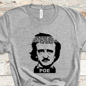 Puede incluir: Una camiseta gris con un gráfico en blanco y negro de Edgar Allan Poe y la palabra "POE" debajo. Encima del retrato están los títulos de sus obras: "El gato negro", "El corazón delator" y "El cuervo".