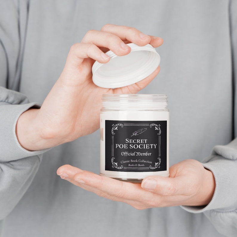 May include: A white candle jar with a black label that reads "Secret Poe Society Official Member Classic Book Collection Books & Bands". The lid is white and is being lifted off the jar.