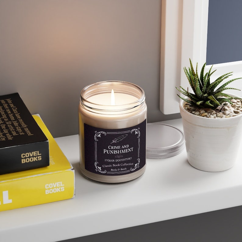 May include: A glass candle jar with a beige label that reads "Crime and Punishment" by Fyodor Dostoevsky. The label is black with white lettering and a decorative border. The candle is lit and the jar is sitting on a white shelf.