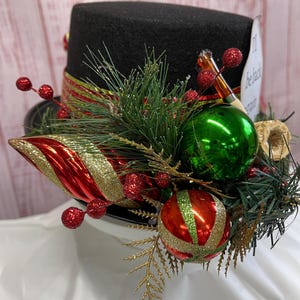 May include: A black top hat decorated with green and gold pine sprigs, red and green striped ornaments, and red berries. The hat has a green and gold striped band and a white sign that says "It's beginning to look a lot like Christmas".