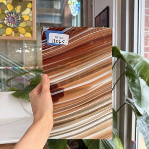 May include: A rectangular piece of glass with a flowing pattern of brown, white, and tan stripes. The dimensions are 11 x 6.5 inches, as indicated by a blue sticker. The glass is held up in front of a window, with natural light illuminating the surface.