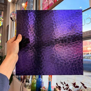Op de afbeelding: Een vierkante plaat van getextureerd, dieppaars glas-in-lood wordt tegen het licht gehouden. Het glas heeft een geribbeld oppervlak, dat het licht opvangt en een glinsterend effect creëert. Het glas is ongeveer 30 cm bij 30 cm.