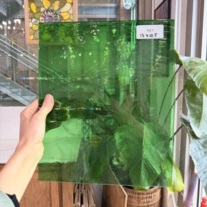 May include: A rectangular piece of transparent green glass is held up, revealing the texture and color. The glass measures 13 x 10.5 inches, as indicated by a white label. The glass is likely for stained glass art or other craft projects.