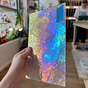 May include: A rectangular sheet of iridescent glass with a textured surface. The glass reflects a spectrum of colours, including blue, yellow, and purple. A blue label with the text "10x6 UM279" is affixed to the top edge. The glass is held in a person's hand.