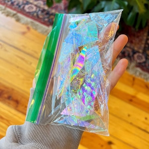 May include: A clear plastic bag filled with iridescent holographic stickers. The stickers are in various shapes and sizes, with a rainbow of colors.