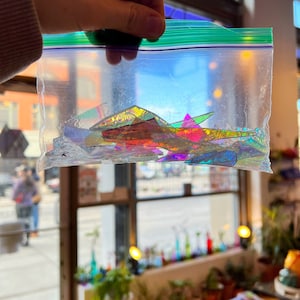 May include: A clear plastic bag filled with iridescent glass shards in various shapes and sizes. The shards are a mix of rainbow colors, including red, orange, yellow, green, blue, and purple.