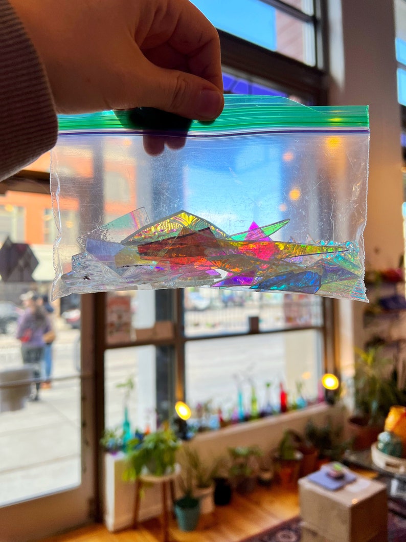 May include: A clear plastic bag filled with iridescent glass shards in various shapes and sizes. The shards are a mix of rainbow colors, including red, orange, yellow, green, blue, and purple.